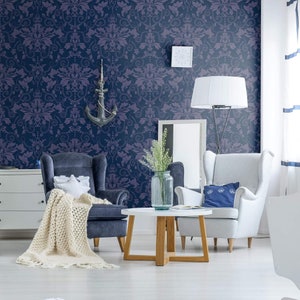 May include: A living room interior with two armchairs, a coffee table, and a white dresser. The walls are covered in a dark blue wallpaper with a light purple damask pattern. The room is decorated with a white lamp, a vase of flowers, and a throw blanket.