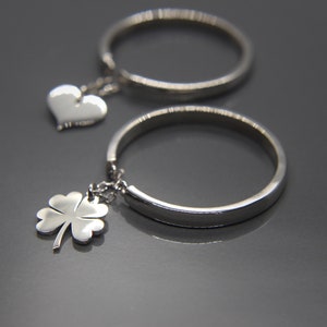 Puede incluir: Dos anillos de plata con dijes. Un anillo tiene un dije de corazón y el otro tiene un dije de trébol de cuatro hojas.