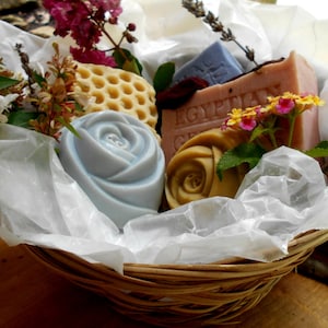 Puede incluir: Una cesta de mimbre llena de una variedad de productos de baño y cuerpo, incluyendo jabones, una esponja vegetal y flores. Los jabones tienen forma de rosas y presentan una variedad de colores, incluyendo azul, rosa y amarillo. La cesta está forrada con papel de seda blanco.