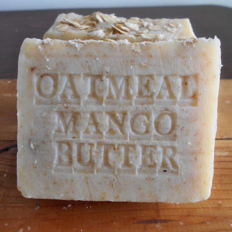 May include: A rectangular bar of soap with the words "Oatmeal Mango Butter" embossed on the front. The soap is light beige with visible flecks and topped with rolled oats. The soap is sitting on a wooden surface.