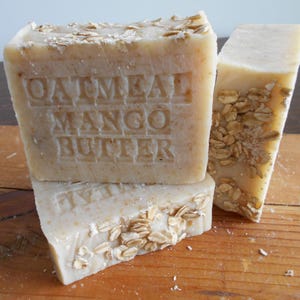 May include: Three bars of handmade soap with the text "Oatmeal Mango Butter" embossed on one. The soap is a light beige color, with visible oatmeal flakes on the surface. The bars are stacked on a wooden surface.