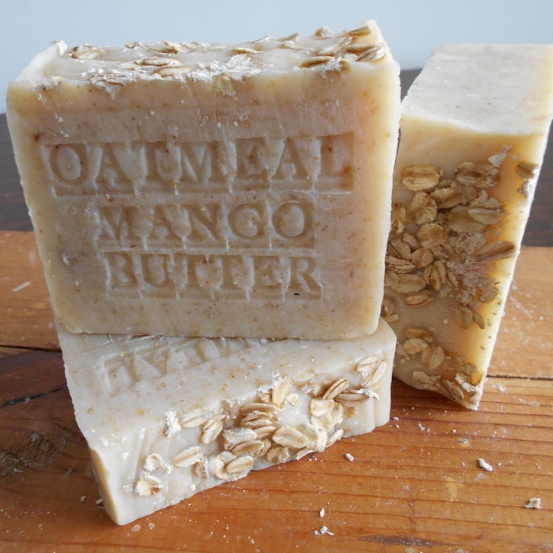 May include: Three bars of handmade soap with the text "Oatmeal Mango Butter" embossed on one. The soap is a light beige color, with visible oatmeal flakes on the surface. The bars are stacked on a wooden surface.