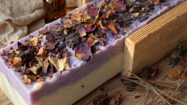 Puede incluir: Un bloque rectangular de jab&oacute;n de dos tonos con una capa base de color blanco crema y una capa superior de color violeta claro. La superficie est&aacute; cubierta con una densa dispersi&oacute;n de p&eacute;talos de flores secas en tonos rosa, rojo y marr&oacute;n.
