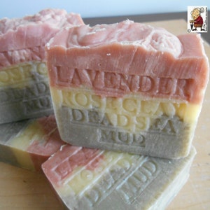 Puede incluir: Una pastilla de jabón rectangular con tres capas horizontales distintas en tonos rosa, amarillo y gris. El texto "LAVENDER", "ROSE CLAY" y "DEAD SEA MUD" está grabado en la superficie del jabón, que tiene un borde superior texturizado.