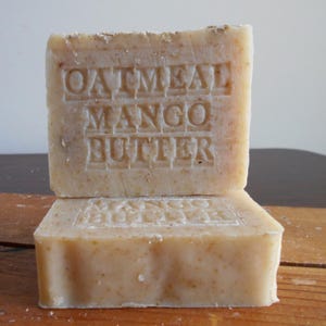 May include: Two rectangular bars of soap stacked on top of each other. The top bar is stamped with the words "OATMEAL MANGO BUTTER" in capital letters. The soap has a light beige color and a textured surface, suggesting natural ingredients.