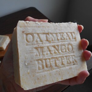 May include: A rectangular bar of handmade soap with the words "OATMEAL MANGO BUTTER" embossed on the surface. The soap is a light beige color and appears to have a slightly rough texture. The soap is held in a person's hand.