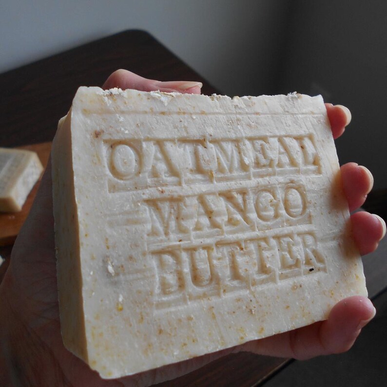 May include: A rectangular bar of handmade soap with the words "OATMEAL MANGO BUTTER" embossed on the surface. The soap is a light beige color and appears to have a slightly rough texture. The soap is held in a person's hand.