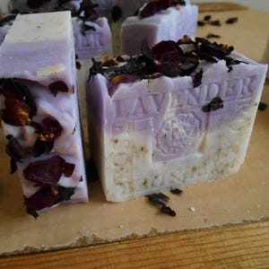 Puede incluir: Barras de jab&oacute;n rectangulares de color lavanda y blanco con p&eacute;talos de flores de color rojo oscuro secos en la parte superior. El jab&oacute;n est&aacute; grabado con el texto LAVENDER y un dise&ntilde;o de sello circular. Las barras tienen una apariencia de color en capas con una parte superior violeta claro y una base color crema, mostrando inclusiones bot&aacute;nicas visibles en toda la textura.