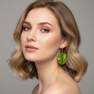 May include: A woman with blonde hair wearing green leaf-shaped earrings with gold-coloured hooks. The earrings are layered, with a detailed vein pattern. The background is a solid, neutral grey.