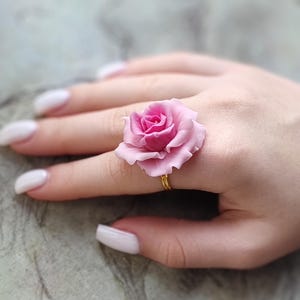 Könnte beinhalten: Ein Ring mit einer rosa Rose und einer goldenen Banderole. Der Ring wird an einem Finger getragen.