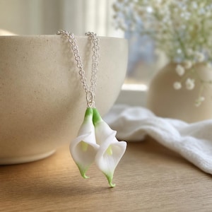 Può includere: Una collana con catena in metallo color argento con un ciondolo a forma di due fiori di calla bianchi con steli verde chiaro. I petali sono lisci e curvi, appesi a una delicata catena a maglie che sostiene il design floreale.