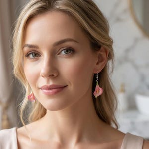 May include: A woman wearing pink calla lily earrings. The earrings are a light pink color with yellow centers and green accents. The earrings are hanging from silver hooks. The woman has blonde hair and is smiling.
