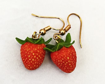 May include: A pair of strawberry earrings. Each earring features a miniature red strawberry with green leaves and gold-colored ear wires. The strawberries have a textured surface with small yellow seeds.