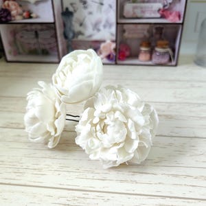 Puede incluir: Cuatro flores de pelo de peonía blanca con horquillas negras. Las flores están agrupadas, mostrando sus pétalos en capas. El fondo presenta una superficie de madera y cajas decorativas.