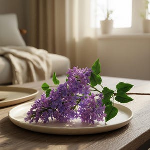 以下が含まれることがあります： 緑の葉が付いた紫色のライラックの花の枝が、クリーム色の皿の上に置かれています。 皿は木製のテーブルの上にあり、背景にはぼやけたソファと窓があります。