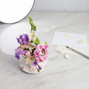 Peut inclure: Une petite tasse à thé remplie d'un arrangement floral avec des iris violets, des pivoines roses et des fleurs blanches. La tasse est posée sur une soucoupe assortie, avec des pétales éparpillés. Un pinceau et une carte vierge sont également présents.