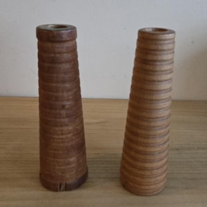 May include: Two wooden spools with a conical shape. The spools are made of a light brown wood and have a series of horizontal ridges. The spools are standing on a wooden surface.