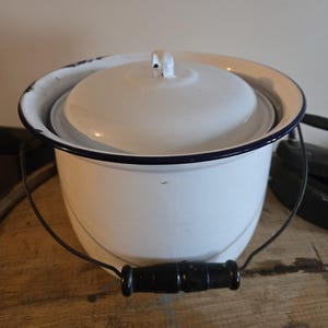 May include: A white enamelware bucket with a lid. The bucket has a black handle and a black rim. The lid has a small handle. The bucket is sitting on a wooden surface.