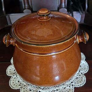 May include: A round, brown ceramic pot with a matching lid and two side handles. The pot features a glossy, speckled glaze finish with darker brown accents along the rim and handle edges, presenting a classic, functional kitchen vessel design.