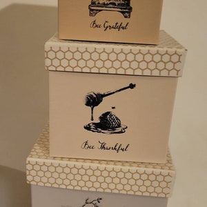 May include: Three stacked decorative boxes with bee-themed illustrations. The boxes are beige, pink, and light gray, each with a different design and the phrases "Bee Grateful," "Bee Thankful," and "Bee Joyful." The lids have a honeycomb pattern.