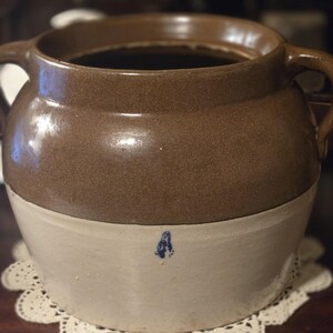 May include: A two-handled stoneware crock featuring a glossy brown glazed upper section and a matte beige lower section. The vessel has a wide circular opening and a small, distinct blue mark stamped on the front of the lower beige portion.