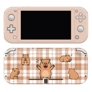 May include: A blush-colored handheld gaming console with a black screen. The back features a brown and white plaid pattern with cartoon capybara illustrations. The capybaras are in various poses, including sleeping and waving.