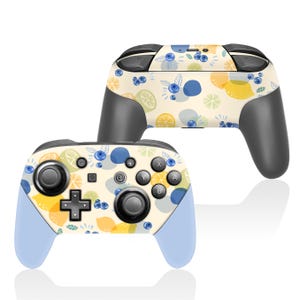 May include: Two wireless game controllers with a fruit pattern. The controllers have a light beige background with lemons, blueberries, and blue circles. The grips are gray, and the bottom of the controllers are light blue.