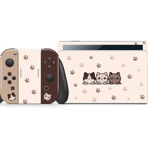 Może przedstawiać: Beżowo-brązowa konsola Nintendo Switch z motywem kota. Kontrolery Joy-Con mają nadruki łapek i ilustracje kotów. Tył konsoli ma pasujący wzór z trzema kreskówkowymi kotami i odciskami łap.