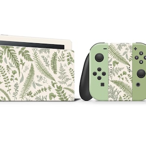以下が含まれることがあります： 植物のシダ模様が施されたNintendo Switchのコンソールとコントローラー。コンソールドックとコントローラーは、クリーム色の背景に薄い緑色です。シダのデザインは、さまざまな緑の色合いを使用しています。