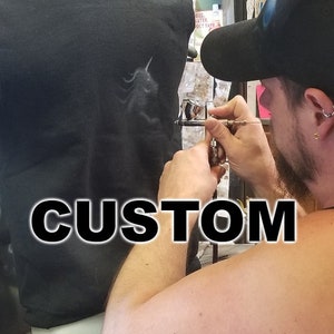 Könnte beinhalten: Eine Person verwendet eine Airbrush, um ein individuelles Design auf einem schwarzen Hoodie zu erstellen. Das Design ist ein weißes Einhorn. Das Wort "CUSTOM" ist in großen, fetten Buchstaben am unteren Rand des Bildes gedruckt.