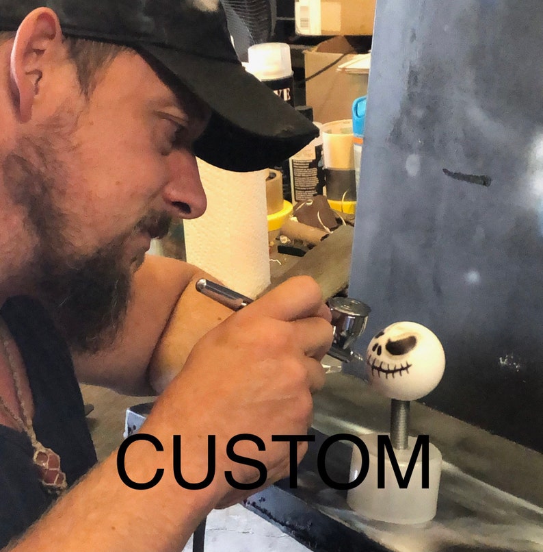 Pode incluir: Uma pessoa est&aacute; usando um aer&oacute;grafo para pintar uma cabe&ccedil;a de cer&acirc;mica branca de Jack Skellington. A cabe&ccedil;a est&aacute; montada em uma base preta com a palavra "CUSTOM" impressa nela.