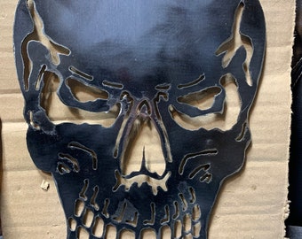 May include: A black metal skull silhouette with cut-out details. The skull features eye sockets, a nose cavity, and a detailed jawline. The metal has a reflective surface. The skull is placed on a cardboard background.