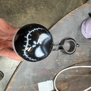 May include: A black steering wheel spinner with a white graphic design. The spinner is attached to a metal clamp. The image is taken from a slightly elevated angle, showing the spinner held in a hand.