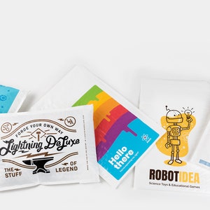 May include: Five colorful, illustrated, paper bags with different designs. The bags feature a robot, a dog, a bird, a cartoon character, and a hammer and anvil. The text on the bags reads "Counter Bot", "Forge Your Own Way Lightning Deluxe", "Hello there", "Robot Idea", and "Slurps Pet Food Treats".