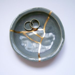 May include: A small, round, grey ceramic bowl featuring a kintsugi-style repair with gold-colored lines running across the surface. Two plain, thin, gold-colored metal rings are resting inside the bowl, which has a smooth, glossy finish.