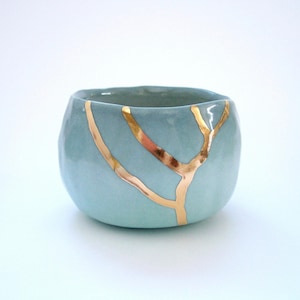 May include: A small, round, light blue ceramic bowl with gold-colored Kintsugi-style repair lines. The bowl has a smooth, glossy finish and a simple, elegant design. The gold lines create a striking contrast against the blue.