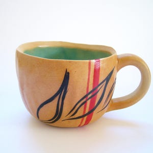 May include: A handmade ceramic mug with a tan exterior and a teal interior. The mug features black and red painted abstract designs. The handle is rounded and the mug has a unique, organic shape. A decorative piece for the kitchen.
