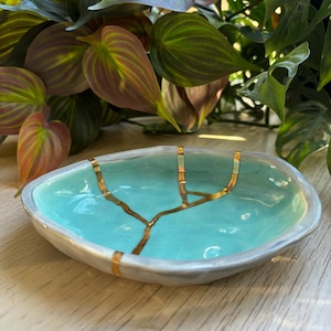 May include: A shallow, irregular ceramic bowl with a glossy turquoise interior and a light gray exterior. The bowl features a prominent gold-colored crack repair design that branches across the center, mimicking the kintsugi technique.