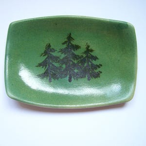 May include: A rectangular ceramic dish with rounded corners, featuring a glossy green glaze and a dark, textured graphic print of three pine trees centered on the interior surface. The dish has slightly raised edges and a smooth finish.