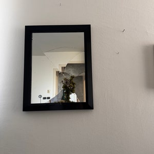 Könnte beinhalten: Ein rechteckiger Spiegel mit schwarzem Rahmen und zerbrochener Glasoberfläche. Der Spiegel ist an einer weißen Wand befestigt. Die Reflexion zeigt einen Raum mit einer Pflanze.