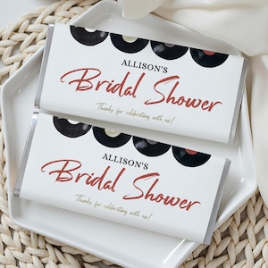 May include: Two white candy bar wrappers with black vinyl record graphics and the text "Allison's Bridal Shower" in red script. The bars rest on a white plate atop a woven mat, with a white cloth draped nearby.