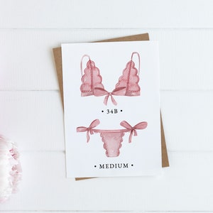 May include: A white card with a watercolor illustration of a pink lingerie set. The card includes the text "34B" and "MEDIUM".