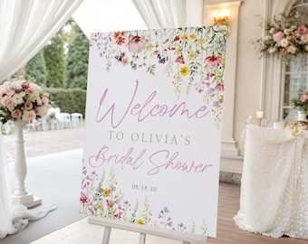May include: A white sign on an easel with the words "Welcome to Olivia's Bridal Shower" in pink script. The sign is decorated with watercolor flowers in pink, purple, yellow, and green. The date "06.18.30" is printed below.