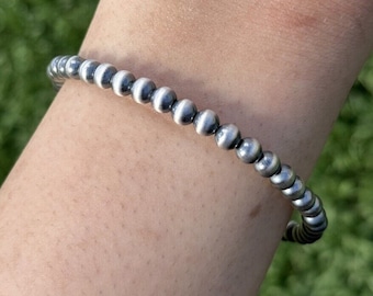 May include: A silver beaded bracelet with a series of small, round beads. The bracelet is worn on a wrist, with a green background visible. The beads have a slightly matte finish.