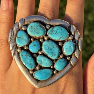 May include: A large, silver-toned ring with a heart-shaped design. The ring is adorned with numerous oval and irregularly shaped turquoise stones. The stones are a vibrant blue-green color, set in individual bezels within the heart frame.