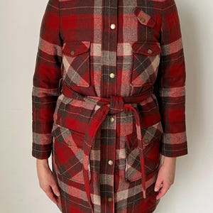 Op de afbeelding: Een rood, bruin en beige geruit jack met een ceintuur in de taille en vier voorzakken. De jas heeft een hoge kraag en wordt gesloten met goudkleurige knopen. De mouwen zijn lang en de jas is gemaakt van warm, geweven materiaal.