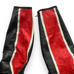 May include: Close-up of black and red leather motorcycle pants. The pants have a central red stripe with white accents and a black stripe on the sides. The leather appears worn, with visible stitching and a zipper.