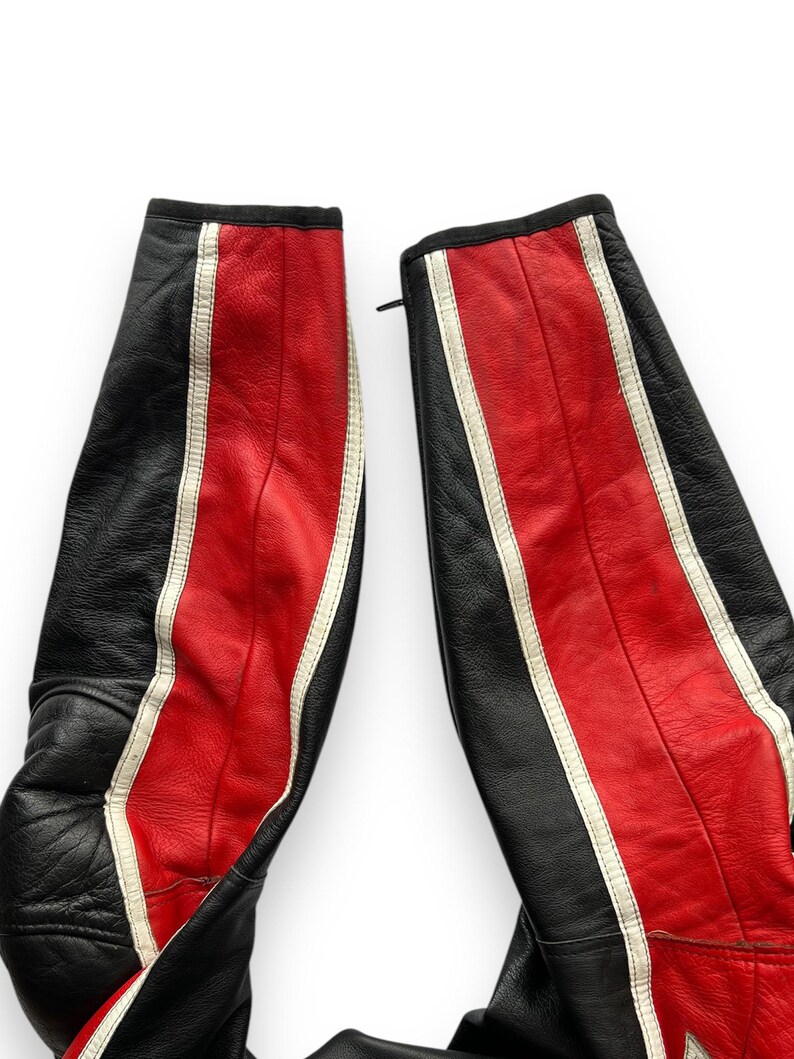 May include: Close-up of black and red leather motorcycle pants. The pants have a central red stripe with white accents and a black stripe on the sides. The leather appears worn, with visible stitching and a zipper.