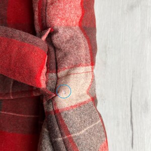 Op de afbeelding: Close-up van een rood en bruin geruite stof, mogelijk een jas of jas. De stof heeft een geweven textuur met een herhalend patroon van vierkanten en rechthoeken. De kleuren zijn een mix van rood, bruin en beige. Het kledingstuk heeft een riem of stropdas.