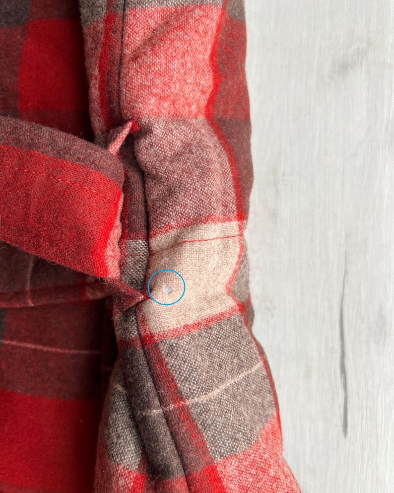 Op de afbeelding: Close-up van een rood en bruin geruite stof, mogelijk een jas of jas. De stof heeft een geweven textuur met een herhalend patroon van vierkanten en rechthoeken. De kleuren zijn een mix van rood, bruin en beige. Het kledingstuk heeft een riem of stropdas.